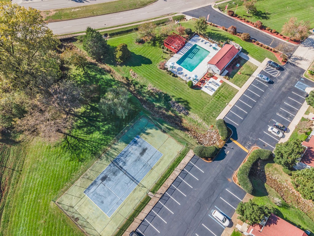 A tennis court is surrounded by a parking lot and a grassy area.