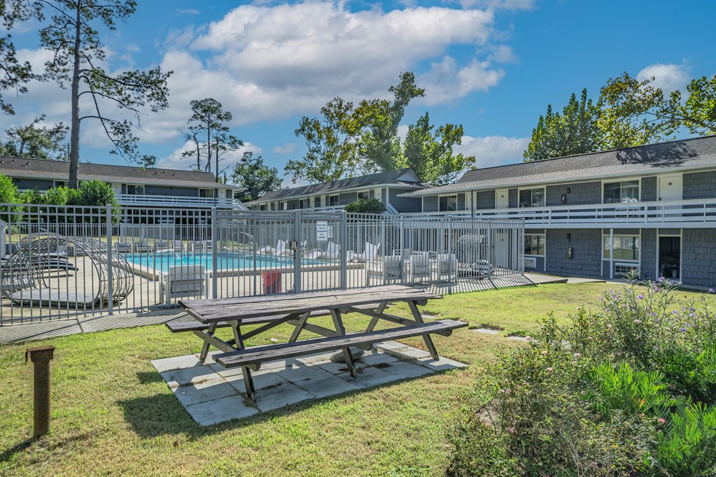 A picnic table sits in the foreground of a sunny outdoor area with a pool and buildings in the background.