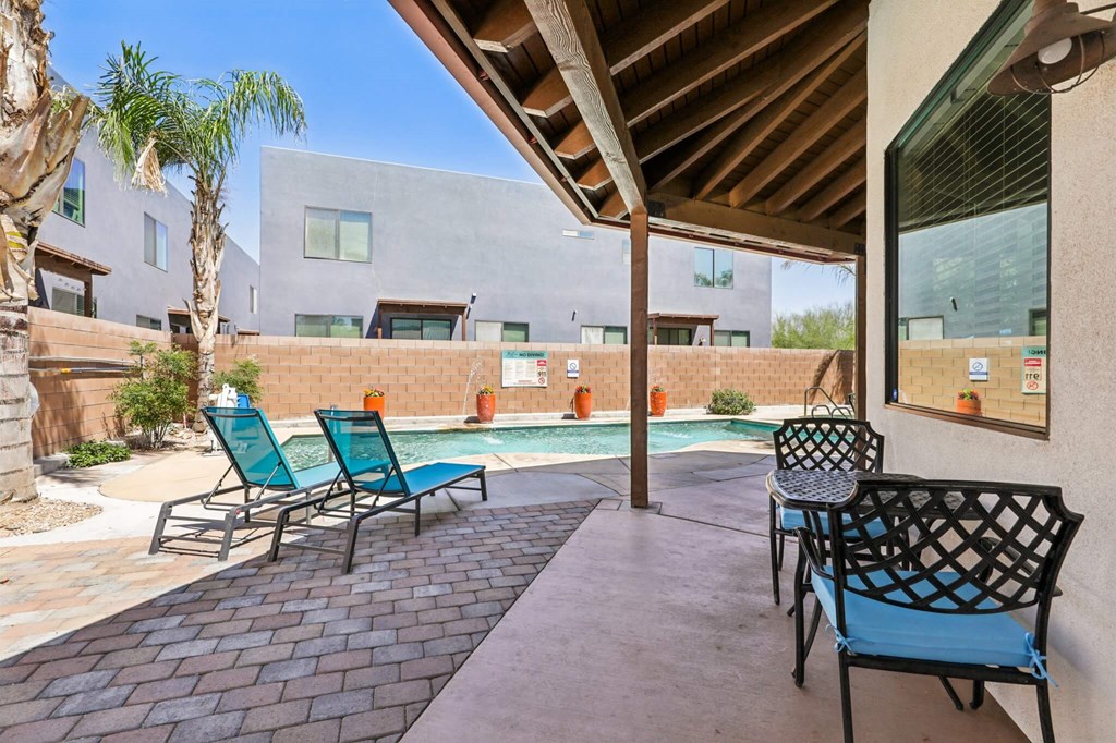 A patio with chairs and a table is surrounded by a pool and buildings.