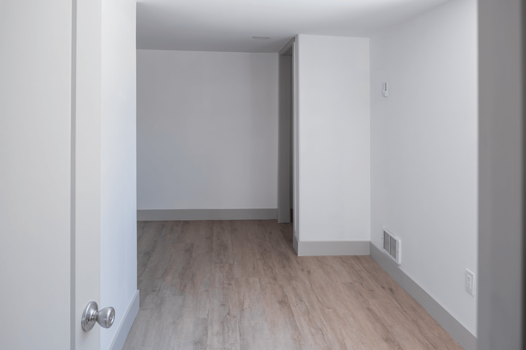 A white door with a silver handle is open to a room with wooden flooring and white walls.