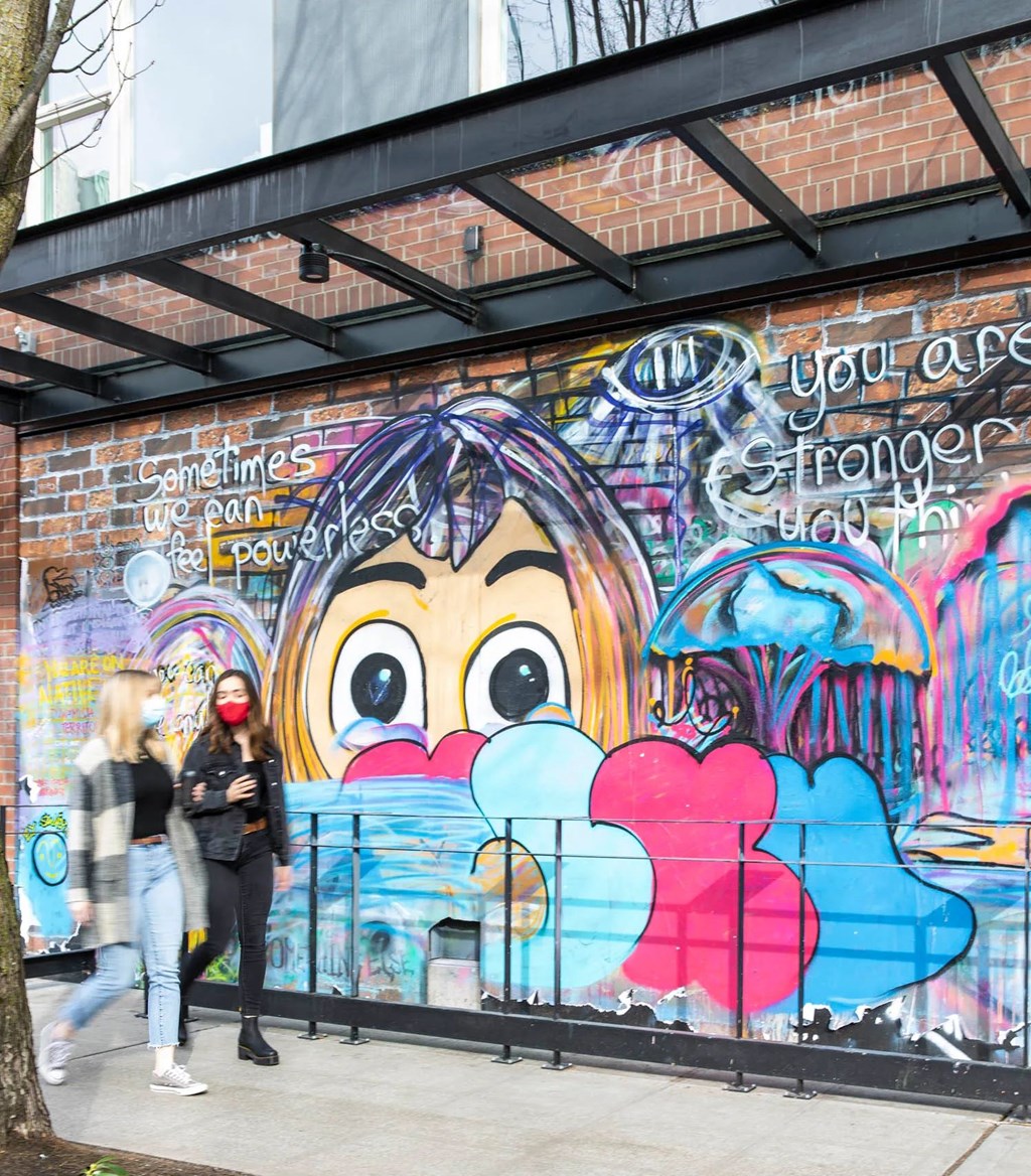 Two women walking past a wall with a mural of a face on it.