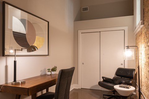 A modern room with a wooden table, a black chair, a black leather chair, a small white table, a plant, a lamp, a painting, a white door, and a stone wall.