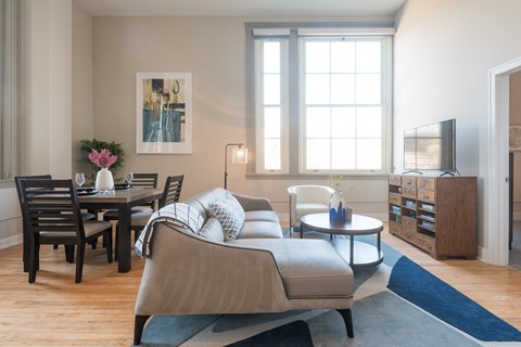 A living room with a grey couch, a wooden dining table with chairs, a coffee table, a television, and a vase with flowers on the table.