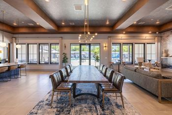 A large wooden dining table with chairs is in the center of a spacious room with a view of the outside.