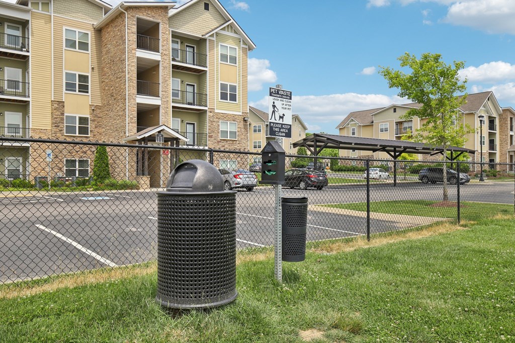 A trash can sits on a grassy area in front of a fence and apartment building at Venue at 109 Apartments, Lebanon , TN, 37090