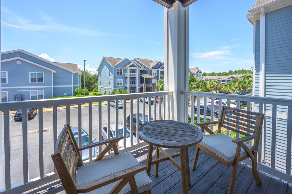 A wooden chair and table are on a porch with a view of a parking lot and houses.