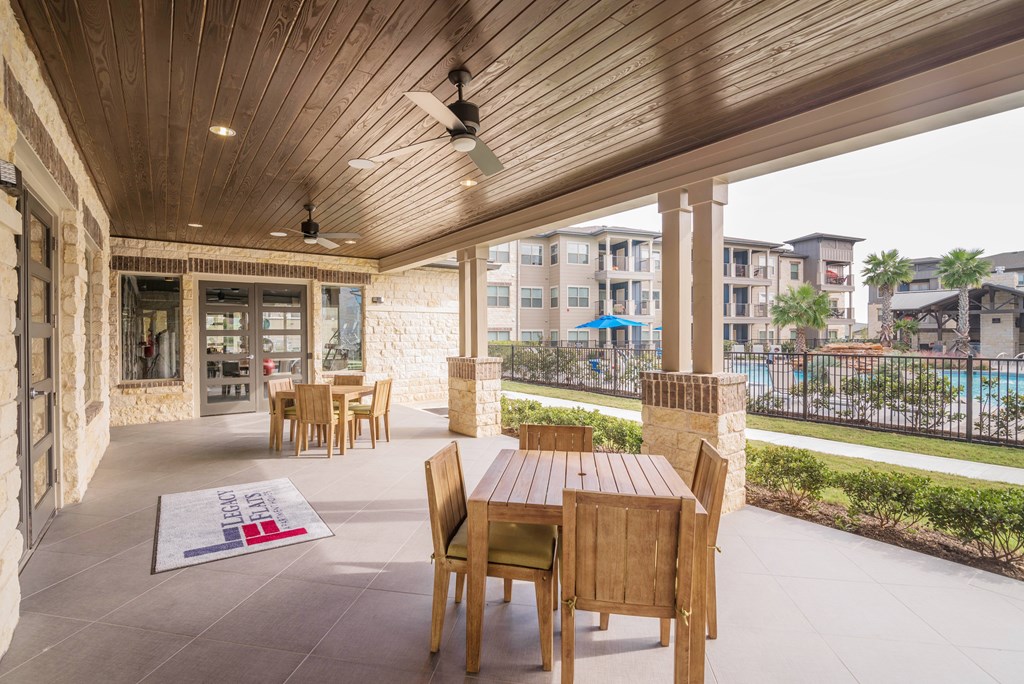 A wooden table and chairs are on a patio with a pool and buildings in the background.