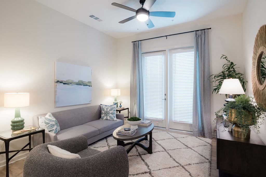 A living room with a grey couch, a grey armchair, a coffee table, a ceiling fan, and a painting on the wall at Hudson at Renaissance Apartments, Wilmington 28405