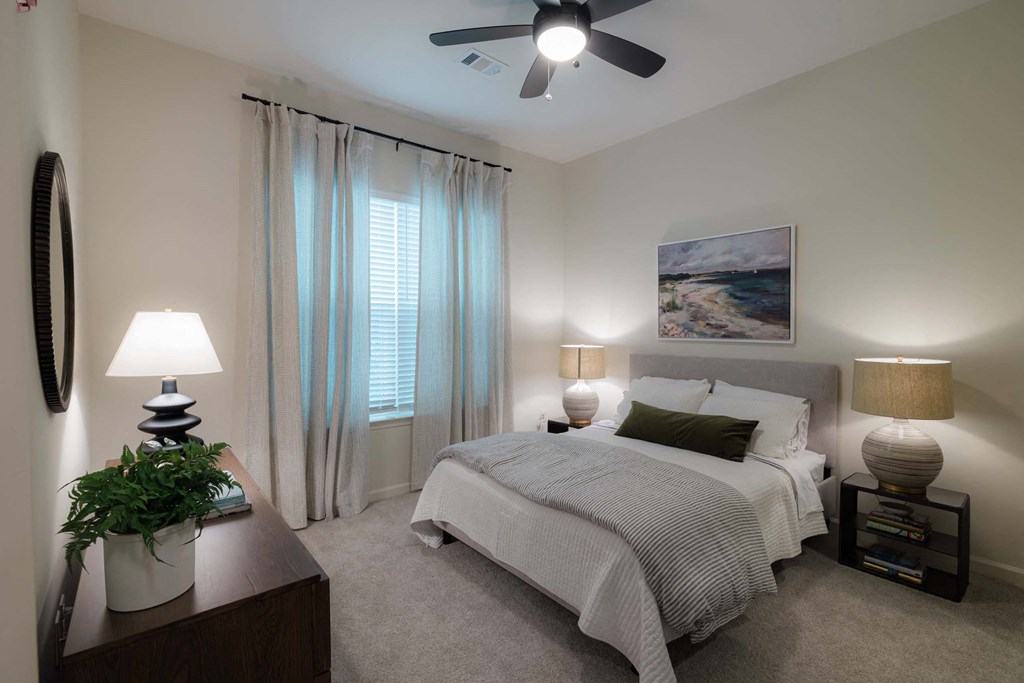A bedroom with a bed, two lamps, a plant, and a painting on the wall at Hudson at Renaissance Apartments, Wilmington, NC 28405