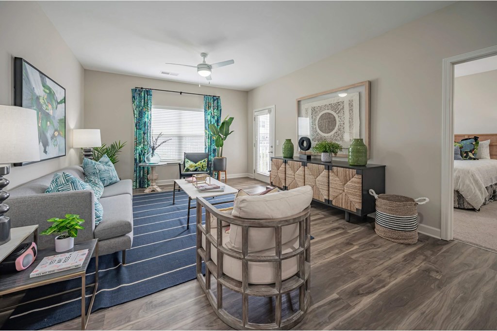 A well-lit living room with a grey sofa, a wooden coffee table, and a bed in the background at Waterleaf at Neely Ferry Apartments, South Carolina 29680