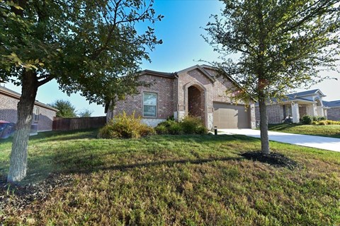 A house with a brown brick exterior and a white garage door is surrounded by trees and grass.