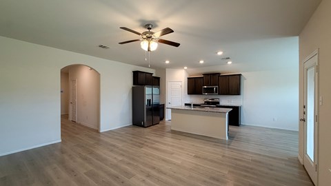 A spacious living room with a ceiling fan and a kitchen area in the background.