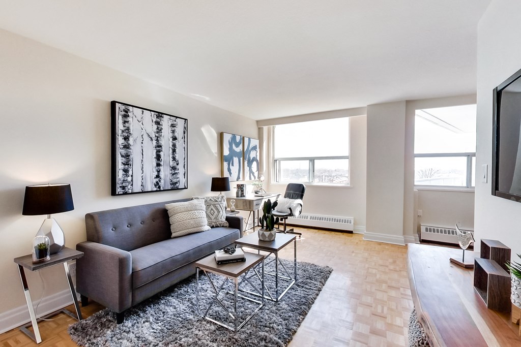 A living room with a grey couch, a black and white picture on the wall, a black floor lamp, a small wooden table with a vase and a plant on it, and a grey area rug.