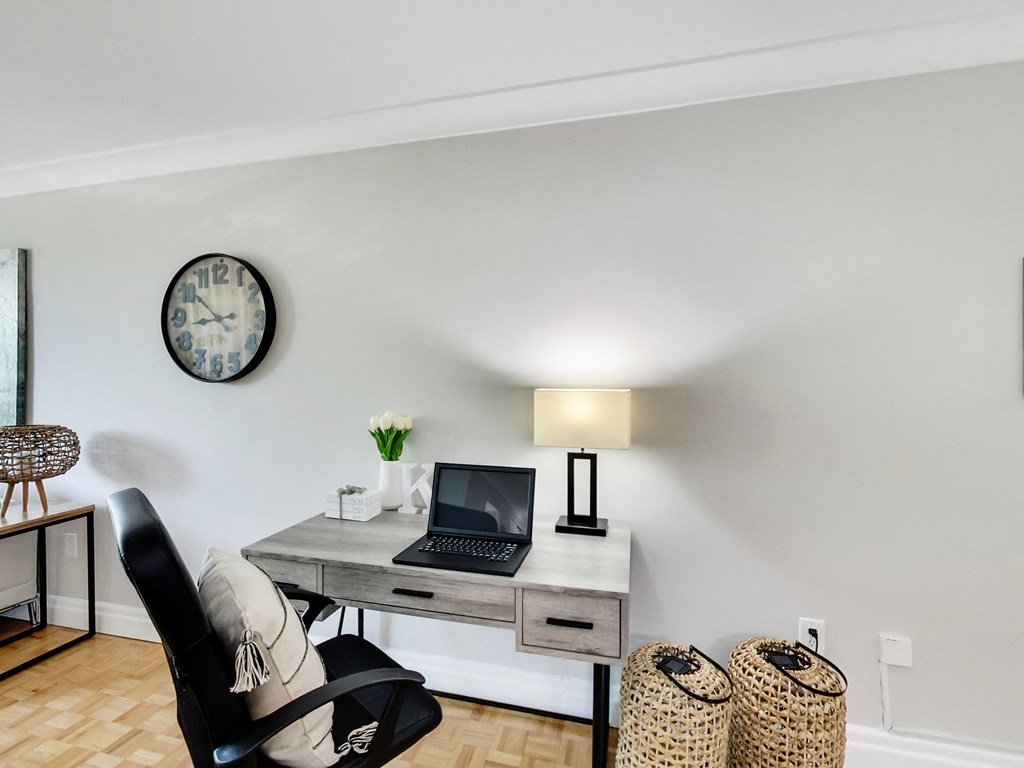 A black chair sits in front of a laptop on a desk with a clock on the wall behind it.