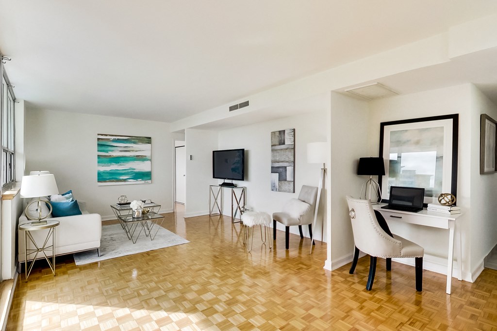 A living room with a white couch, a white chair, a black chair, a white desk, a television, a laptop, a lamp, a painting, and a floor lamp.
