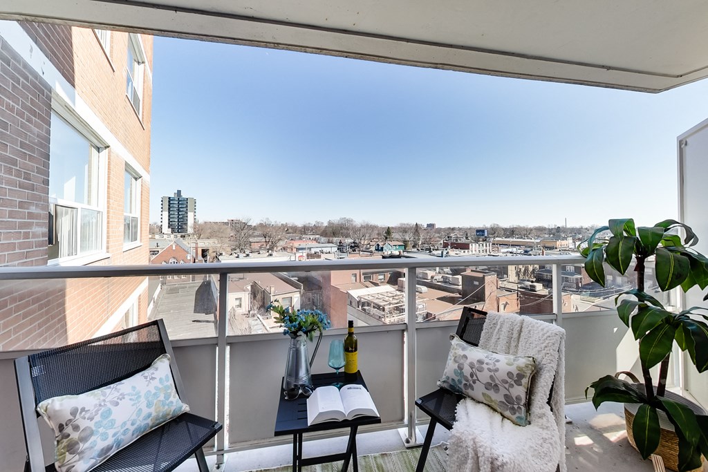 A balcony with a black chair, a black cushion, a black pillow, a white cushion, a white blanket, a green plant, a vase with flowers, a bottle, a book, and a