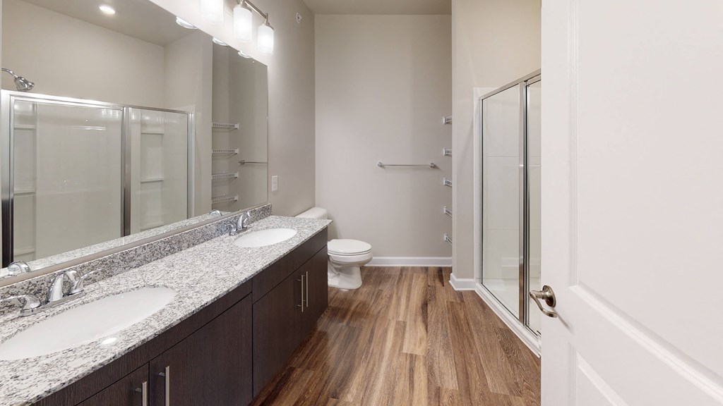 Bathroom with dual sinks, dark cabinets, toilet and shower; vinyl plank floors