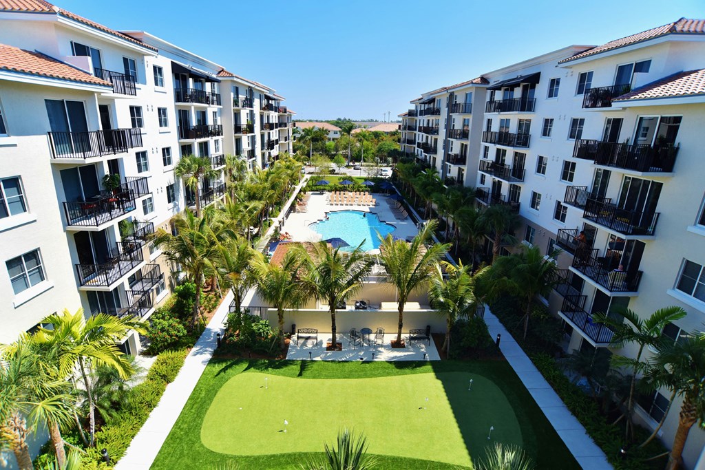 A view of an apartment complex with a pool surrounded by palm trees and a green lawn.