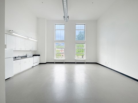 A large empty room with a kitchen area to the left and a window with a view of the outside.