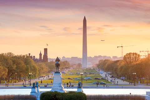 The Washington Monument stands tall in the distance behind a park with a statue on a pedestal in the foreground.