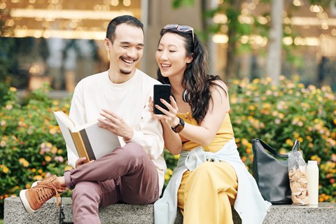 A man and woman are sitting on a bench with a book and a phone in their hands.