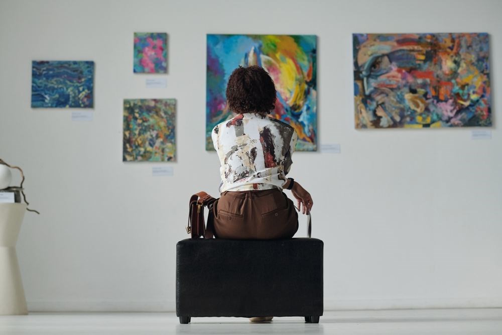 A woman is sitting on a suitcase looking at paintings on the wall.