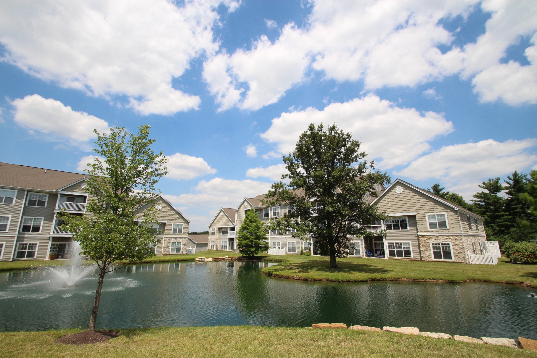 This is a picture of a fountain in a pond with building exteriors at Nantucket Apartments, in Loveland, OH.