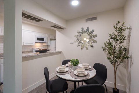 A dining area with a table set for four and a potted plant with a view of the kitchen pass-through window in a 1 bedroom, 1 bath 554 sq ft apartment home at The Biltmore Apartments located in the Vickery Midtown neighborhood of Dallas, TX.
