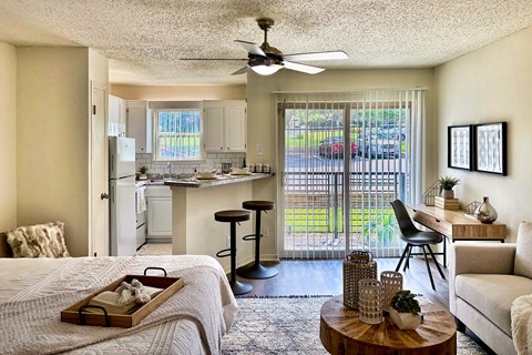 Living and sleeping area in an Efficiency apartment at Cambridge Court Apartments in Dallas, Texas, showing carpeted flooring, patio door, dining space, and connection to the kitchen.