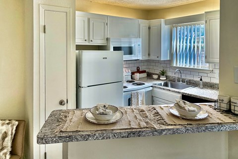 Kitchen and breakfast bar in an Efficiency apartment at Cambridge Court Apartments in Dallas, Texas, showing counter space, cabinetry, appliances, and open connection to the living area.