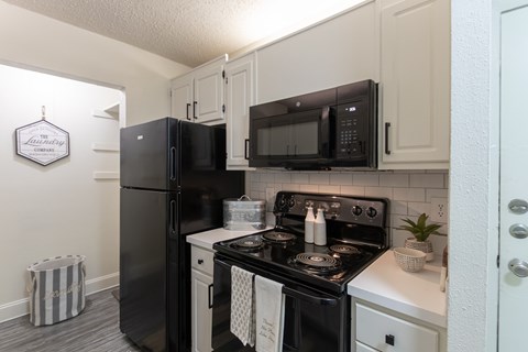 Kitchen view in a 1-bedroom apartment at Cambridge Court Apartments in Dallas, Texas, showing white cabinetry, black appliances, range, microwave, and pantry area.