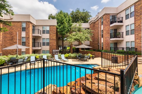 A swimming pool with a waterfall surrounded by lounge chairs and a couple of tables, chairs and umbrellas at Cambridge Court Apartments Entryway in Lake Highlands, Dallas, TX.