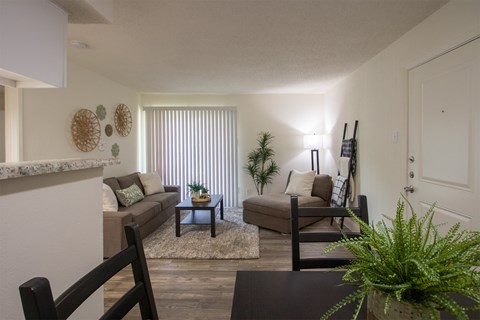 View of the living room with a couch, chaise lounge and a potted plant from the dining room of a 515 square foot 1 bedroom, 1 bath apartment home at Canyon Creek Apartments in Dallas Midtown neighborhood of Dallas, TX.