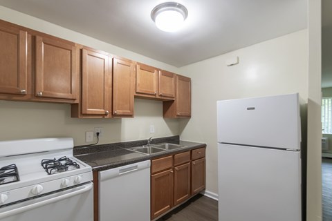 Another angle of the Colonial Ridge kitchen, with a double-basin sink, dishwasher, wood cabinetry, and white appliances arranged along one wall.