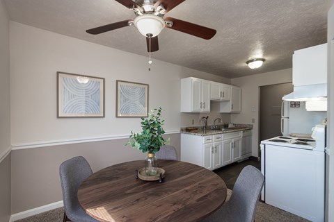 Dining area in the Fairfield Pointe 1-bedroom apartment, with a round table, ceiling fan, framed wall art, and kitchen in the background.