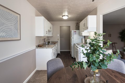 Dining area beside the kitchen in the Fairfield Pointe 1-bedroom model, with a round table, white cabinetry, and kitchen walkway visible.