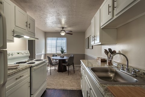 Galley kitchen in the Fairfield Pointe 850 sq. ft. 1-bedroom apartment, with white cabinets, sink, range, refrigerator, and dining area beyond.