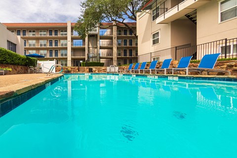 A pool area with lounge chairs, a patio table and chairs with a blue umbrella and stainless steel BBQ in front of apartment buildings at Princeton Court Apartments in the Vickery Midtown neighborhood of Dallas, TX.