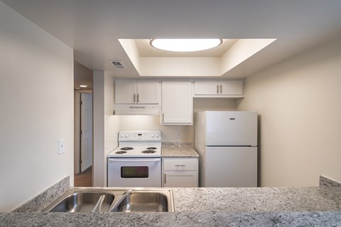 Kitchen counter view at Village East Apartments, looking across the double sink toward white cabinets, refrigerator, and electric range.