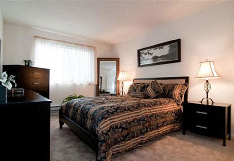 A bedroom with a patterned bedspread, a bed with a headboard, a nightstand with a lamp, and a dresser with a mirror.