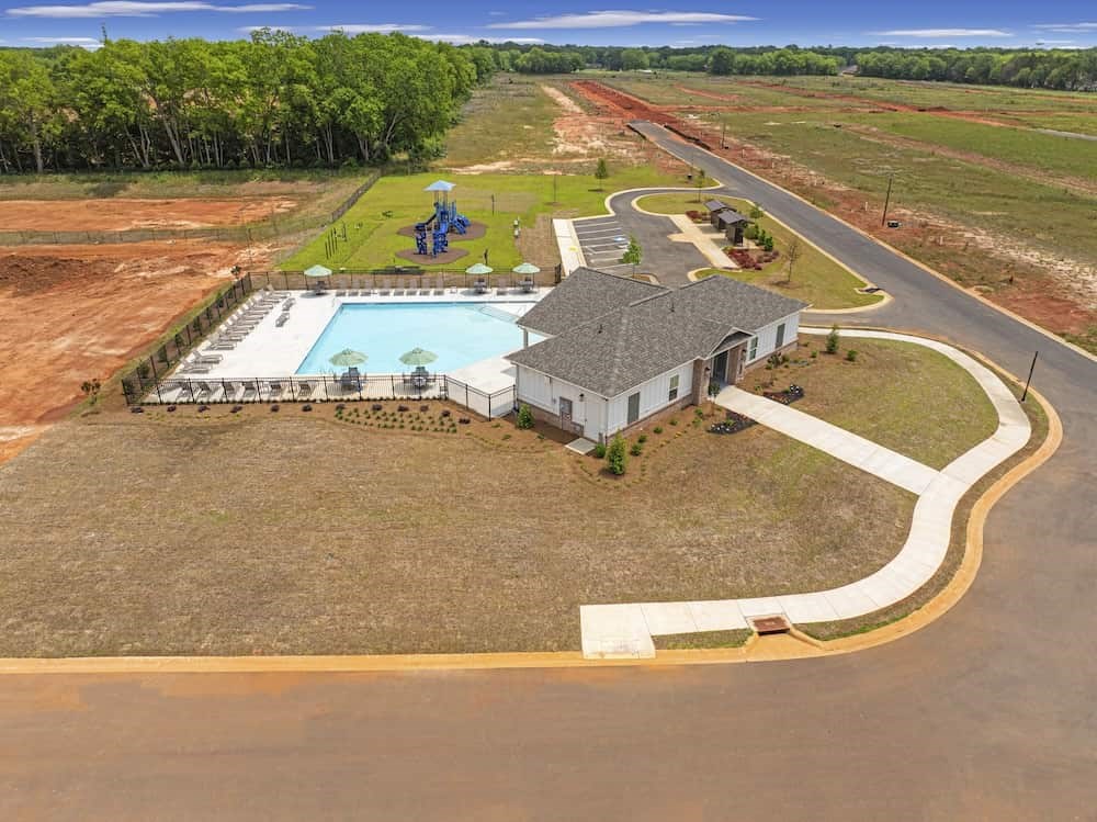 A house with a pool and a playground is surrounded by a grassy area and a dirt field.