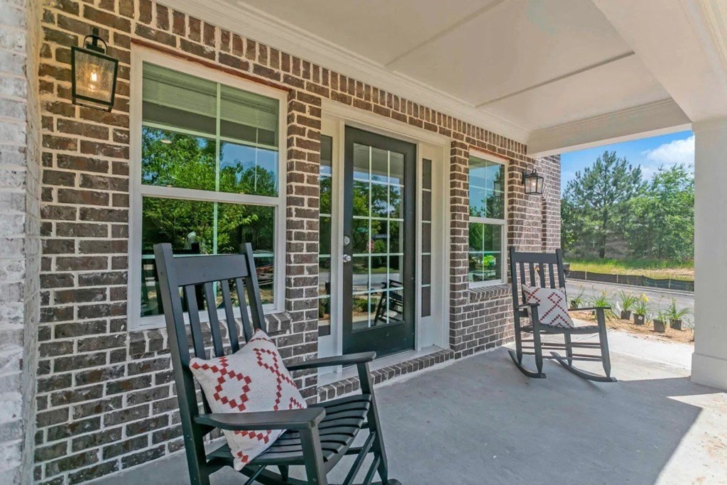 Two chairs are on a porch with a brick wall and a window.