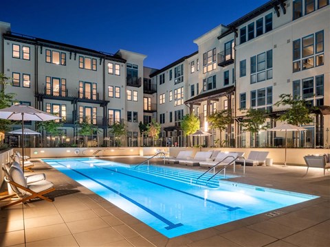 A swimming pool surrounded by lounge chairs and umbrellas in front of apartment buildings.