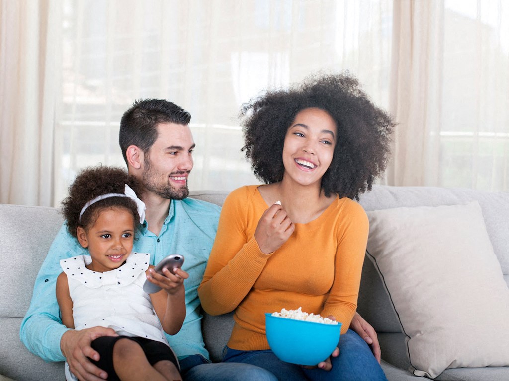 A family of three is sitting on a couch, with the woman holding a bowl of popcorn and the girl holding a remote control.