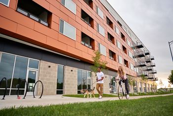 A woman riding a bicycle next to a man walking a dog in front of a modern building.at Linc, Des Moines, IA  