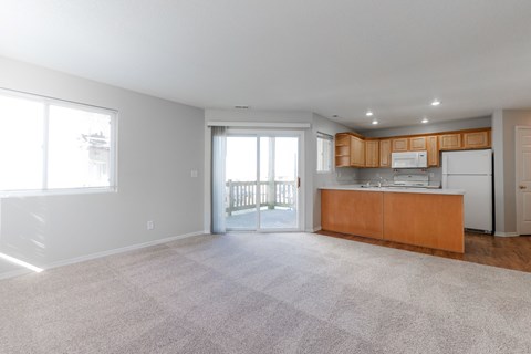 A spacious living room with a carpeted floor and a kitchen area with wooden cabinets and a white refrigerator.