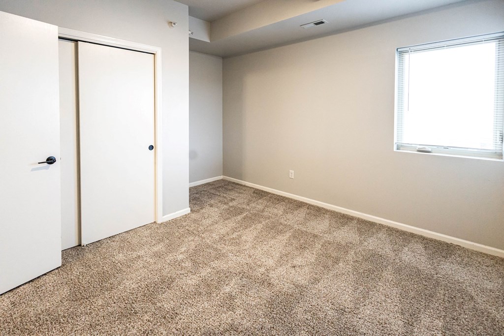 A room with a carpeted floor, a white door, and a window with blinds at Meadowlark Place Apartments, Grimes