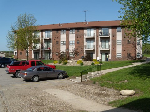 A grey car is parked in front of a red truck and a brown building.