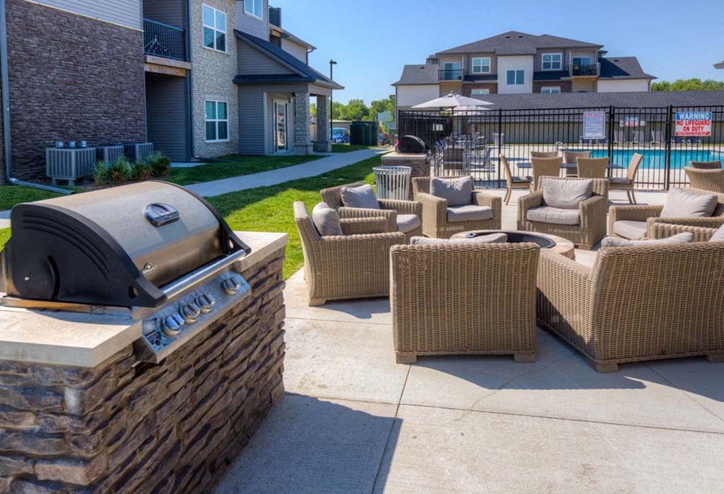 A barbecue grill is on a stone wall next to a patio with wicker chairs. at Stonegate Crossing Apartments, Clive, IA, 50325