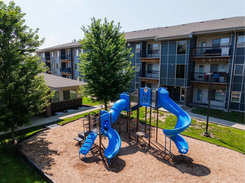 A playground with a blue slide in the foreground and apartment buildings in the background.